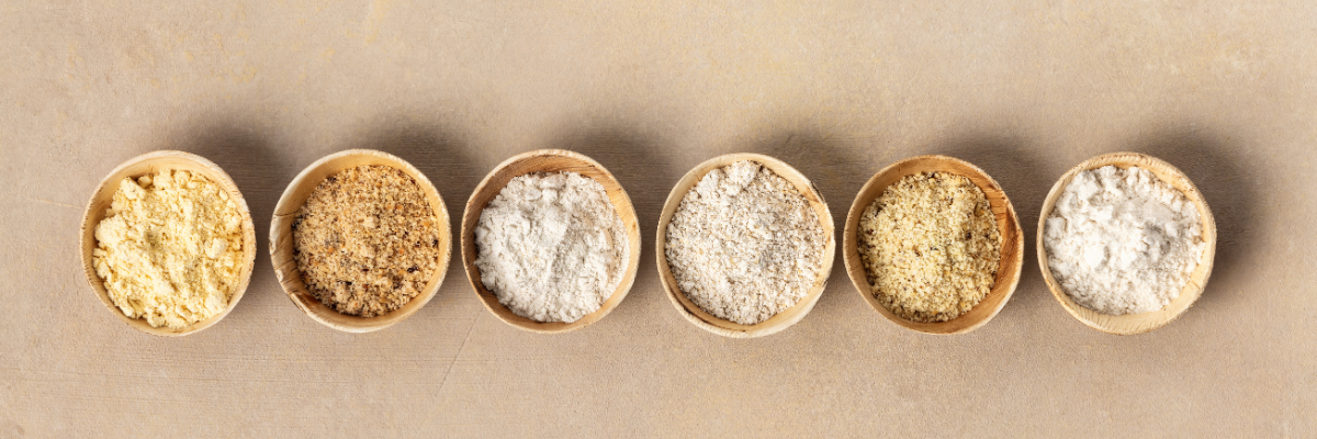 Six bowls of various flour on a natural surface.