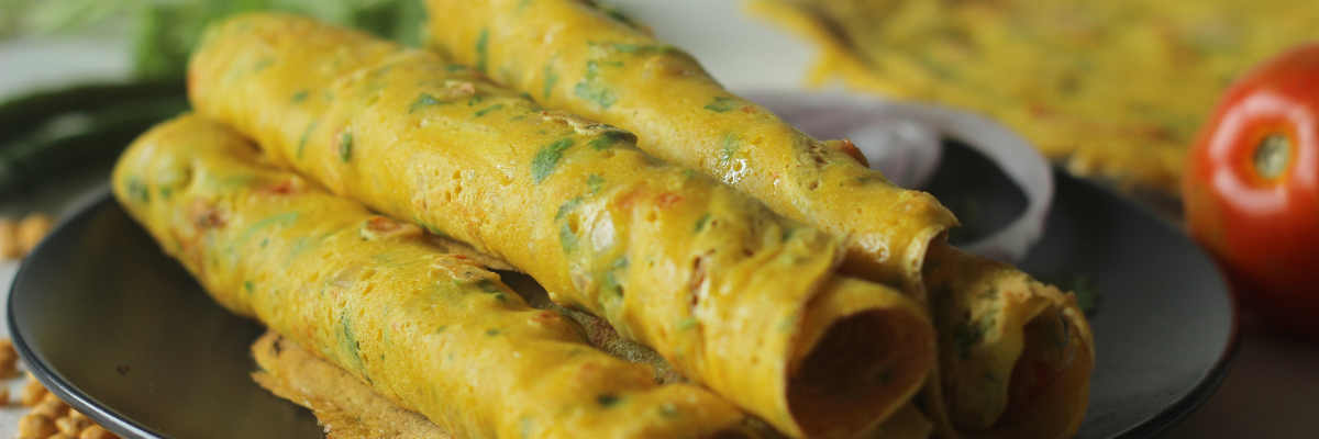 A stack of rolled chilla on a stone plate with various ingredients in a blurred background. 
