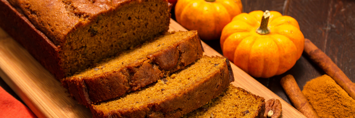 Prebiotic Pumpkin Spice Banana Bread on a wooden surface with spices and couple of small pumpkins nearby.