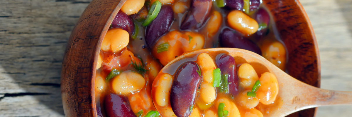 A wooden bowl of veggie baked beans with a wooden spoon on a pale wood surface.
