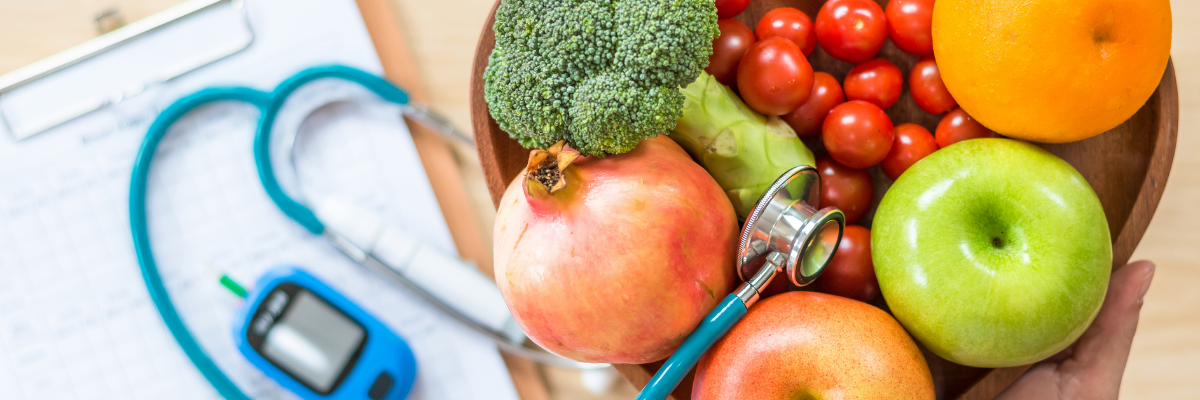 A heart-shaped wooden bowl of fresh produce hovers over medical paperwork and a glucose testing device.