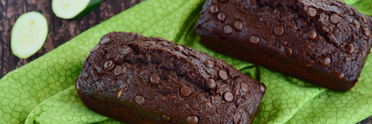 Two loaves of chocolate-chocolate chip zucchini bread, with slices of zucchini nearby, atop a green cloth on a dark wooden surface. 