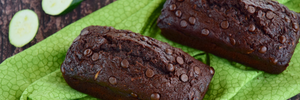 Two loaves of chocolate-chocolate chip zucchini bread, with slices of zucchini nearby, atop a green cloth on a dark wooden surface. 