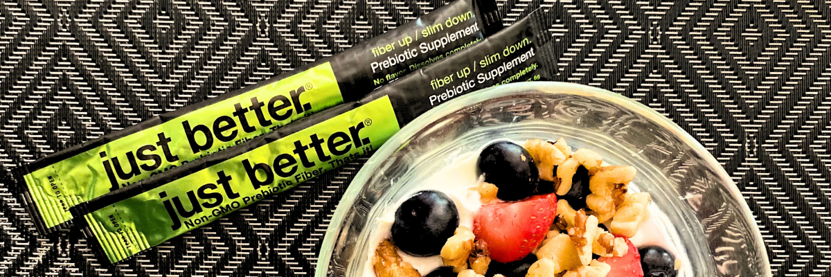 A gray and black patterned background with two stick packs of just better fiber with a bowl of fruit, nuts, and yogurt. 