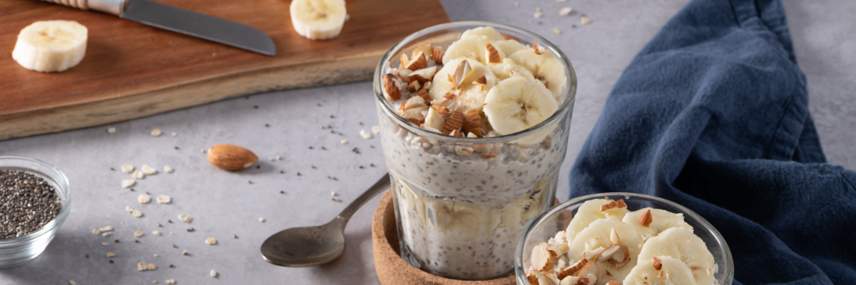 Two servings of Banana Chia Overnight Oats with Almonds are on a countertop, and sliced bananas are on a cutting board nearby. 
