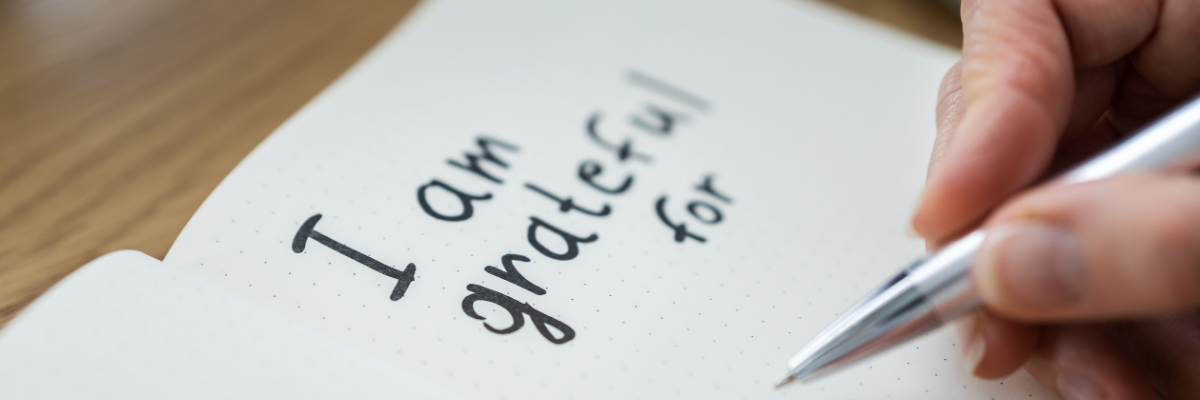 A hand writes I am grateful for on a notebook with a pen.