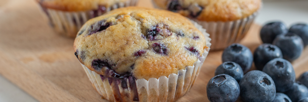 Several freshly baked blueberry muffins on a wooden surface with fresh scattered blueberries nearby. 