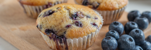 Several freshly baked blueberry muffins on a wooden surface with fresh scattered blueberries nearby. 