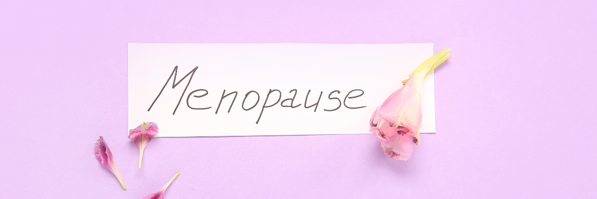 A pink background with a piece of paper that reads: Menopause. Pink flower petals are scattered around the paper.