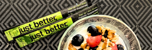 A gray and black patterned background with two stick packs of just better fiber with a bowl of fruit, nuts, and yogurt. 