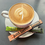 Hot coffee drink with latte art on a saucer with a stick pack of just better prebiotic fiber and a stick pack of just better allulose 