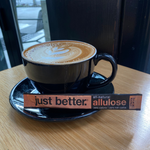 Cappuccino in a black cup with a bar of 'just better' allulose on a wooden table.